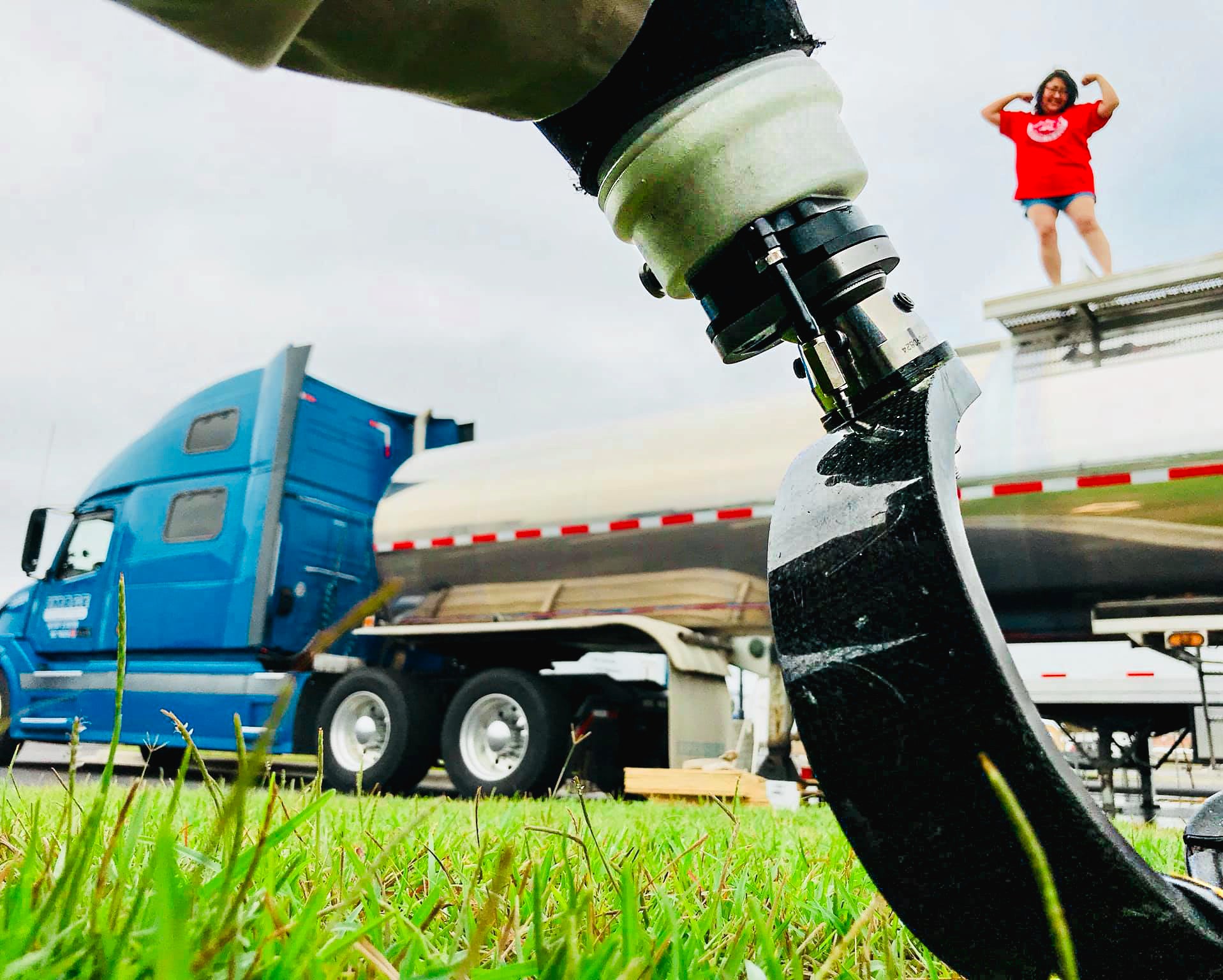 Detail photo of an Amplife® Supporter's prosthetic leg with their companion standing on top of 18 wheeler flexing in the background